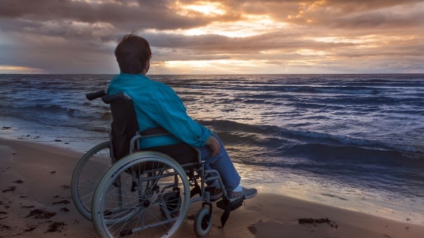 Social theme A disable woman in a wheelchair looks at a sunset on the beach by the sea Her folding wheelchair is part of her life serving as her legs Evening Dark key